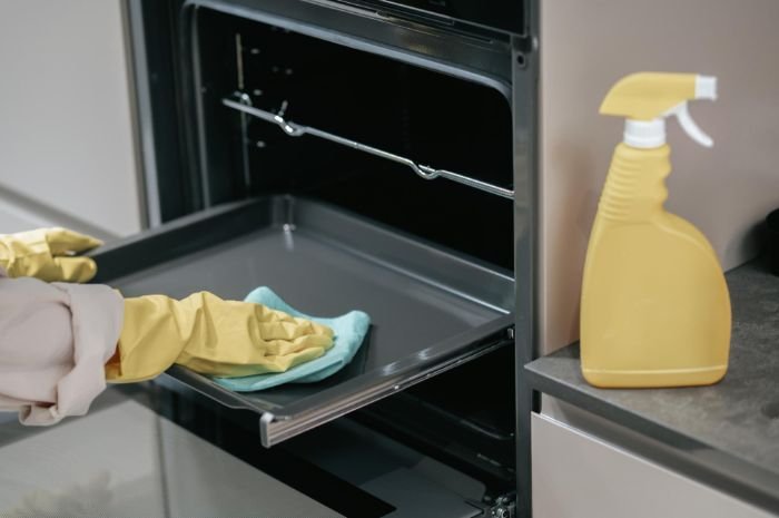 Close-up of hands wearing yellow gloves wiping the oven door with a microfiber cloth next to a spray bottle, illustrating a step in the oven cleaning guide.