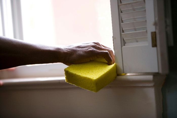 A person's hand wiping a windowsill with a yellow sponge, representing the pre-cleaning steps necessary for effective pest control in a home.