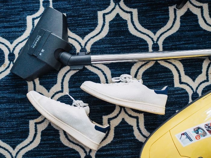 Vacuum cleaner head on a patterned carpet with sneakers aside, depicting a quick clean-up in a bedroom setting.