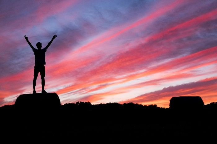 The silhouette of a person with arms raised in victory stands on a rock against a dramatic sunset sky, symbolizing the achievement of reaching cleaning goals during the move-out process.