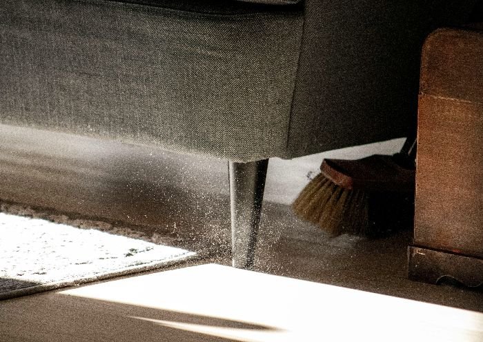 Close-up of dust being swept from under a sofa, revealing the often-neglected areas that require cleaning for a meticulous bedroom upkeep.