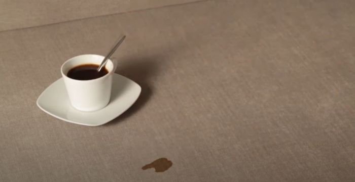 A cup of coffee on a saucer with a small spill on a taupe upholstered surface, highlighting the risk of stains on furniture.