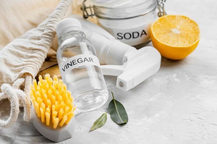 A bottle of vinegar, a jar of baking soda, and a citrus fruit next to a scrub brush, representing a natural cleaning solution for kitchen appliances.