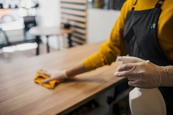 A person in a yellow shirt and black apron cleaning a wooden counter with a cloth and spray bottle, representing the efficiency and thoroughness of professional cleaning services.